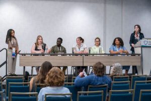 Five panelists sit at a long table on a small stage, passing a microphone around as they answer questions for the audience.